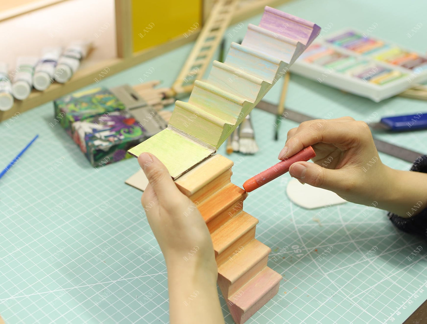 Hands painting wooden iLAND miniature dollhouse stairs, showcasing adjustable design with vibrant colors on a crafting table.