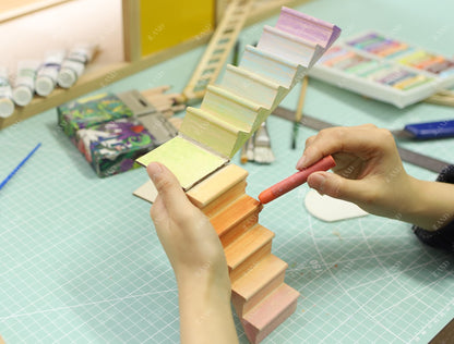 Hands painting wooden iLAND miniature dollhouse stairs, showcasing adjustable design with vibrant colors on a crafting table.